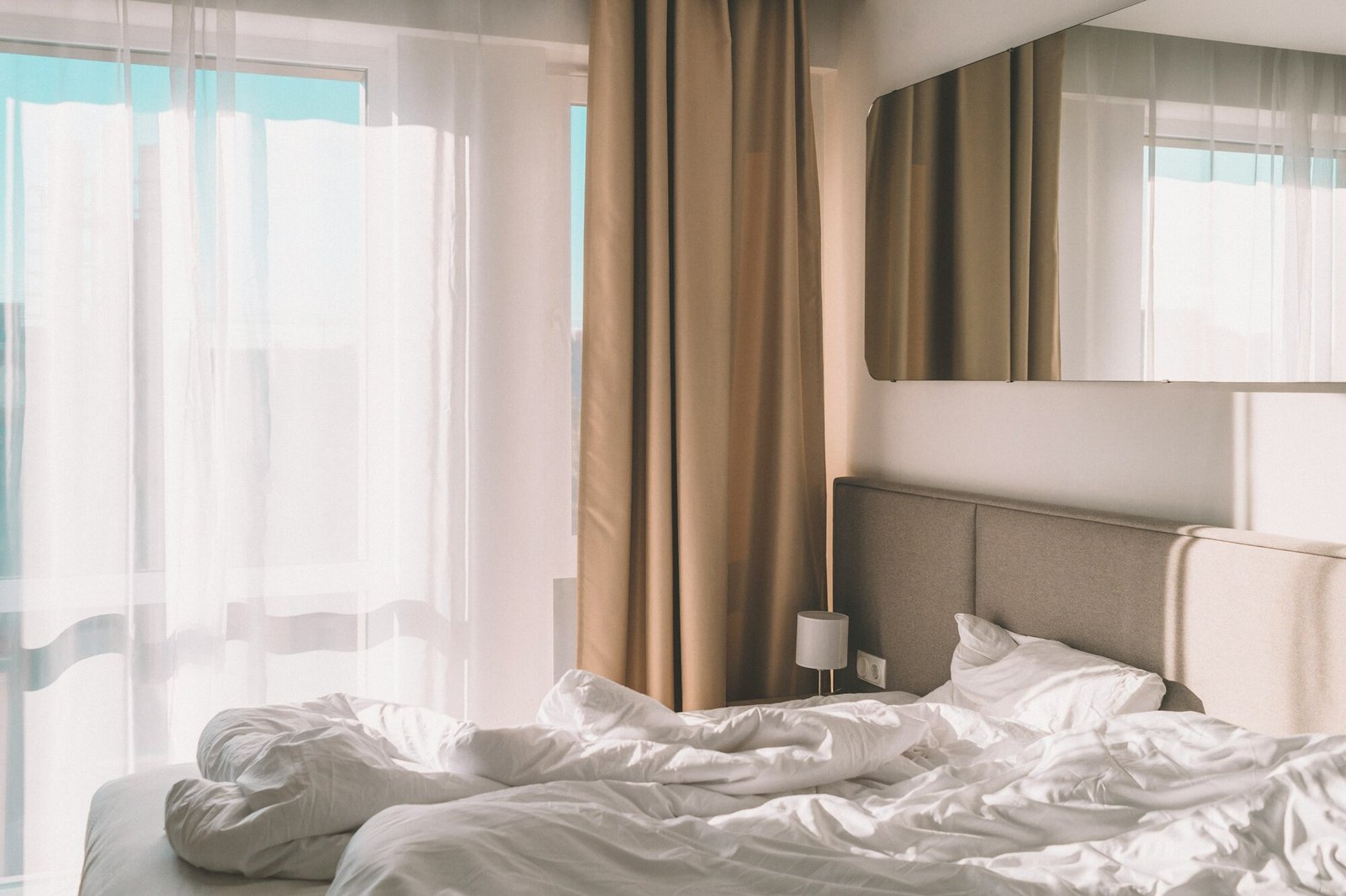 Bright bedroom with white linen bedding, sheer curtains, and warm morning daylight.