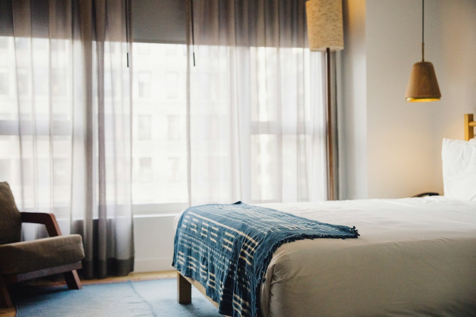 Boutique hotel bedroom with sheer curtains, a soft armchair, and a textured throw on the bed.